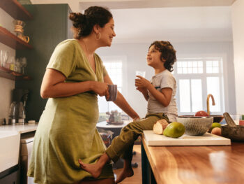 Happy mother and child enjoying a calm morning in a bright, modern kitchen. The mother leans on the counter with a coffee mug while her son sits nearby with a glass of water, surrounded by fresh fruit and natural light — representing family comfort, cleanliness, and pest-free living.